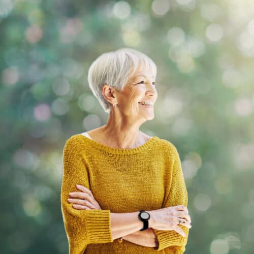 L'image montre une femme senior rayonnante, vêtue d'un pull jaune vif, qui arbore un grand sourire tout en regardant vers l'extérieur. Elle porte un bracelet de téléassistance moderne au poignet, qui est à peine visible mais essentiel pour sa sécurité. Le bracelet souligne la capacité de Tavie Assist à intégrer la sécurité dans le quotidien des seniors sans entraver leur style de vie ou leur indépendance. Le contexte verdoyant et lumineux ajoute une touche de sérénité et de bien-être à l'image, mettant en avant le confort et la tranquillité d'esprit que ces dispositifs apportent à leurs utilisateurs.