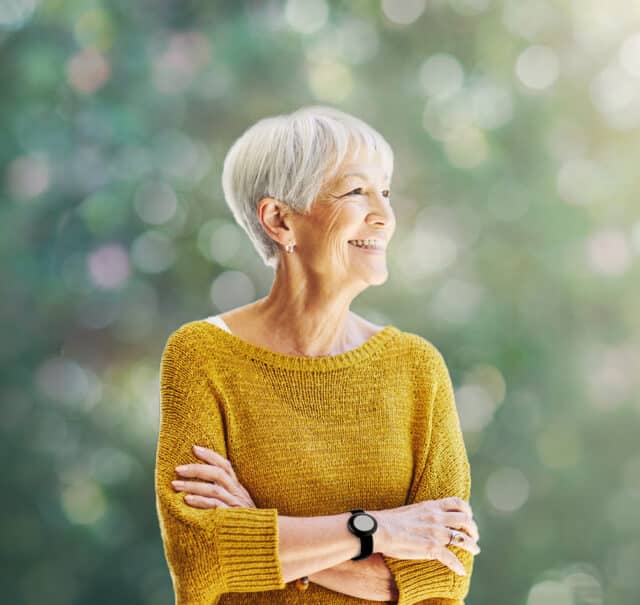 L'image montre une femme senior rayonnante, vêtue d'un pull jaune vif, qui arbore un grand sourire tout en regardant vers l'extérieur. Elle porte un bracelet de téléassistance moderne au poignet, qui est à peine visible mais essentiel pour sa sécurité. Le bracelet souligne la capacité de Tavie Assist à intégrer la sécurité dans le quotidien des seniors sans entraver leur style de vie ou leur indépendance. Le contexte verdoyant et lumineux ajoute une touche de sérénité et de bien-être à l'image, mettant en avant le confort et la tranquillité d'esprit que ces dispositifs apportent à leurs utilisateurs.