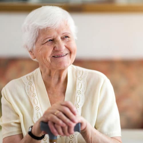 L'image montre une femme âgée souriante, assise confortablement dans son salon. Elle porte un dispositif de téléassistance moderne au poignet, bien que le focus soit mis sur une téléassistance à domicile qui n'exige pas préalablement un bracelet. Cette technologie avancée permet aux seniors de vivre en toute sécurité et autonomie dans leur propre maison, tout en ayant accès à une aide rapide en cas d'urgence. Les solutions de téléassistance sans bracelet utilisent souvent des capteurs environnementaux ou des appareils connectés pour surveiller le bien-être de l'utilisateur, offrant ainsi une sécurité discrète sans nécessiter de porter un dispositif physique.