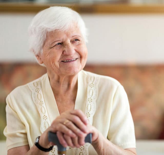 L'image montre une femme âgée souriante, assise confortablement dans son salon. Elle porte un dispositif de téléassistance moderne au poignet, bien que le focus soit mis sur une téléassistance à domicile qui n'exige pas préalablement un bracelet. Cette technologie avancée permet aux seniors de vivre en toute sécurité et autonomie dans leur propre maison, tout en ayant accès à une aide rapide en cas d'urgence. Les solutions de téléassistance sans bracelet utilisent souvent des capteurs environnementaux ou des appareils connectés pour surveiller le bien-être de l'utilisateur, offrant ainsi une sécurité discrète sans nécessiter de porter un dispositif physique.