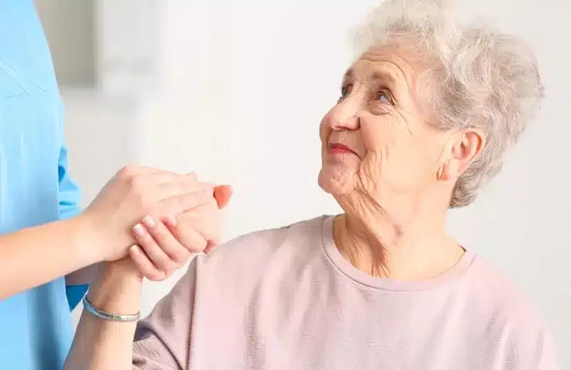 Dans cette image, on voit une femme âgée, les cheveux gris bouclés, levant les yeux vers une personne en tenue de soin bleue qui lui tient doucement la main. L’expression bienveillante de la femme âgée et le geste rassurant de la soignante illustrent la qualité de l’attention et des soins apportés. La communication non verbale entre elles est empreinte de confiance et d'assurance, suggérant que la personne âgée se sent soutenue et respectée. C’est une représentation de la qualité de service et des relations humaines que l’on cherche à établir dans le domaine des soins aux seniors.