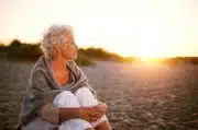 Sur cette image, nous voyons une femme aux cheveux bouclés blancs, assise seule sur une plage au coucher du soleil. Elle est vêtue d’un haut léger et d’un châle, et ses yeux sont fixés au loin sur l’horizon où le soleil s’abaisse, baignant la scène dans une lumière chaude et dorée. Cet instant de calme et de sérénité capture la beauté tranquille d’un moment de réflexion ou de méditation, évoquant la paix intérieure.