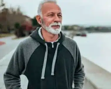  Un homme d’âge mûr, aux cheveux poivre et sel et à la barbe soigneusement taillée, se tient dehors, vêtu d'un sweat à capuche gris. Le sérieux de son regard, tourné vers le lointain, évoque une réflexion profonde. Peut-être pense-t-il à ses souvenirs, à ses expériences, ou bien contemple-t-il simplement le paysage devant lui. Cet homme pourrait symboliser les bénéficiaires de services comme Tavie Alzheimer, qui aide les personnes atteintes de troubles cognitifs à mener une vie plus autonome et sécurisée grâce à la technologie. Derrière lui, l'environnement est paisible, avec une étendue d’eau et un ciel couvert, renforçant l'atmosphère de tranquillité et de soutien que Tavie cherche à offrir.