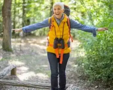 Dans un élan de liberté et d’aventure, cette dame aux cheveux argentés exprime sa joie en pleine nature. Équipée d’un sac à dos et vêtue d’une veste bleue sur un gilet jaune vibrant, elle étend les bras comme pour embrasser l’espace qui l’entoure. Les jumelles autour de son cou et le sourire radieux traduisent son plaisir d’explorer. Grâce à la téléassistance extérieure, elle profite de ces moments précieux avec l’assurance qu’en cas de besoin, l’aide n’est jamais loin, lui permettant de savourer sa randonnée en toute sécurité au cœur de la forêt qui l’entoure.