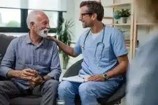 L’image illustre un homme âgé en conversation avec un professionnel de la santé, probablement un infirmier ou un médecin, vu sa tenue bleue de travail. Ils semblent être dans un cadre intime, probablement le salon de la maison de l’homme, ce qui reflète une approche personnalisée et humaine des soins. Cette scène dépeint l'étape 3 d'un service d'urgence où, après avoir été contactés, les opérateurs du service de téléassistance prennent l’initiative d’alerter l’entourage du patient ou de contacter les secours pour assurer l’intervention nécessaire.