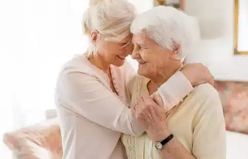  L’image capture un moment tendre de proximité entre deux femmes âgées, qui pourraient être des amies ou des membres de la famille. Elles se tiennent dans une étreinte chaleureuse, l’une souriant doucement à l’autre. La présence de rires et de sourires communique un sentiment de confort et de soutien mutuel. Cette affection témoigne de l’importance des liens personnels et de la proximité dans le bien-être des seniors, un élément central dans l’approche de soins et d’assistance à domicile. 