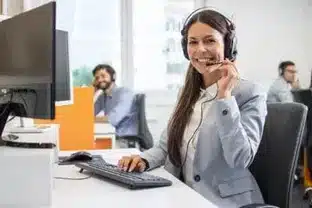 Sur cette image, on voit une opératrice souriante, équipée d'un casque téléphonique, assise devant un ordinateur dans ce qui semble être un centre d'appels. Avec son doigt sur le microphone, geste typique indiquant une communication en cours, elle représente l'étape 2 d'un service d'assistance client : la réponse personnalisée. Son attitude professionnelle et son sourire chaleureux incarnent l'engagement du service à fournir une assistance rapide et adaptée à chaque appelant. En arrière-plan, d'autres opérateurs sont visibles, suggérant une équipe dédiée à répondre aux besoins des clients en continu.