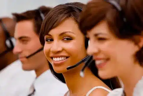 L’image montre une rangée d’opérateurs de centre d’appels, tous équipés de casques téléphoniques. Au premier plan, une jeune femme regarde l’objectif avec un sourire confiant et accueillant. Son attitude professionnelle et son sourire sincère reflètent l'engagement de l’équipe à fournir une qualité de service client irréprochable. Les collègues flous en arrière-plan suggèrent un environnement de travail dynamique et axé sur le travail d'équipe, où chaque membre contribue à l’excellence collective du service proposé aux clients.