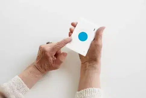 Téléalarme mobile. L’image montre les mains ridées d'une personne âgée tenant fermement un dispositif d'alerte mobile blanc avec un large bouton bleu au centre. Ce dispositif est  L’image montre les mains ridées d'une personne âgée tenant fermement un dispositif d'alerte mobile blanc avec un large bouton bleu au centre. Ce téléalarme mobile est destiné à être utilisé en cas d'urgence pour assurer la sécurité de l’utilisateur. En pressant ce bouton, la personne peut rapidement demander de l'aide, ce qui est essentiel pour les seniors vivant seuls ou ceux qui sont susceptibles de nécessiter une assistance immédiate en cas de chute ou de malaise. L'image évoque la tranquillité d'esprit que de tels dispositifs procurent, tant pour les utilisateurs que pour leurs proches, symbolisant une bouée de sécurité dans le quotidien des personnes vulnérables. vulnérables.