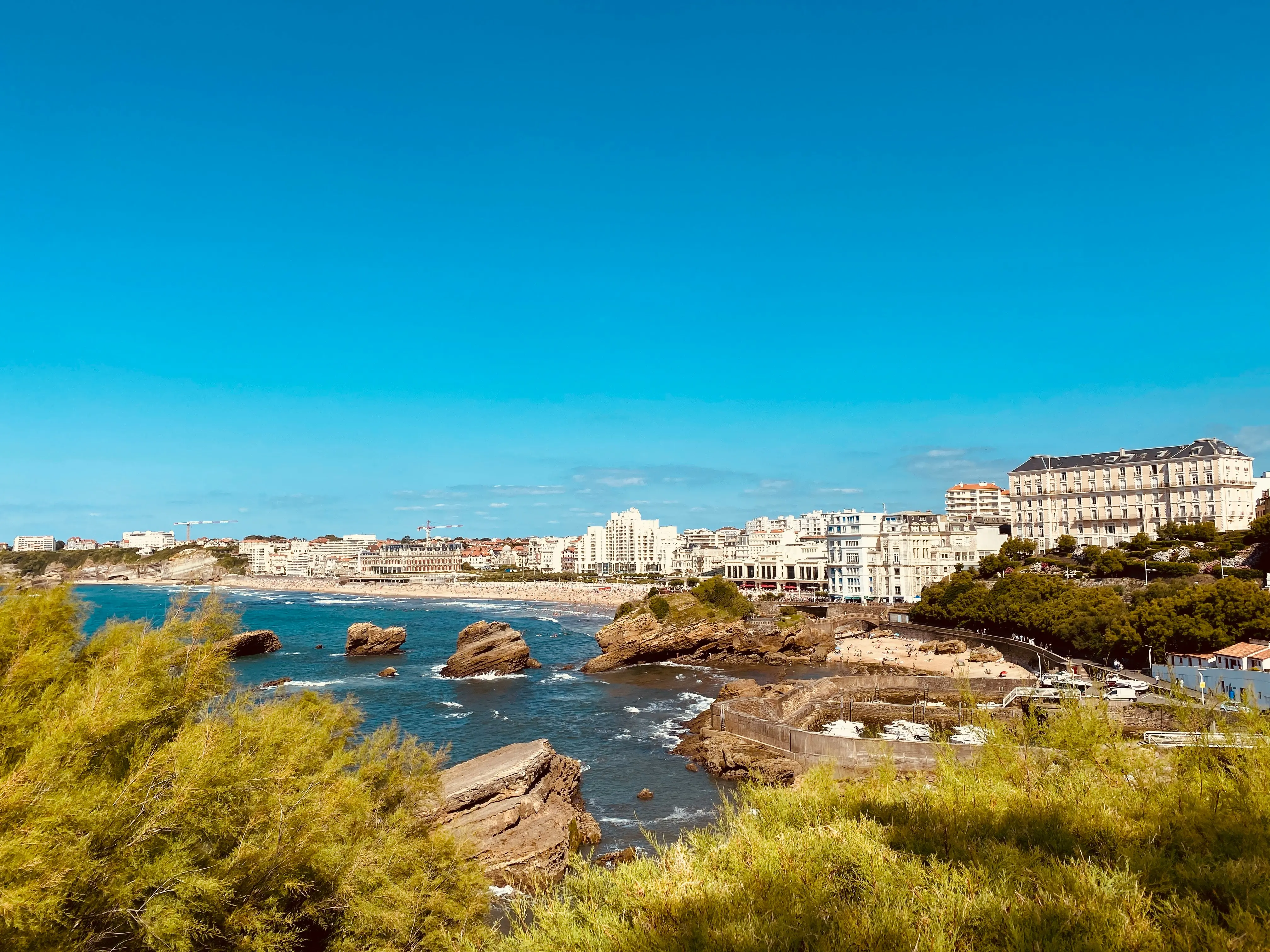 L’image montre un magnifique paysage côtier de Biarritz avec une mer bleu profond, des rochers éparpillés dans l’eau et une ville élégante en arrière-plan sous un ciel dégagé. La végétation verdoyante au premier plan contraste avec les bâtiments blancs et beiges typiques de la région. À Biarritz, la téléalarme assure sécurité et sérénité aux habitants, notamment aux personnes âgées vivant dans ce cadre idyllique.